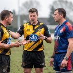 Un jeune arbitre de rugby en tenue officielle explique une décision, sifflet à la bouche, à deux capitaines sur un terrain amateur.